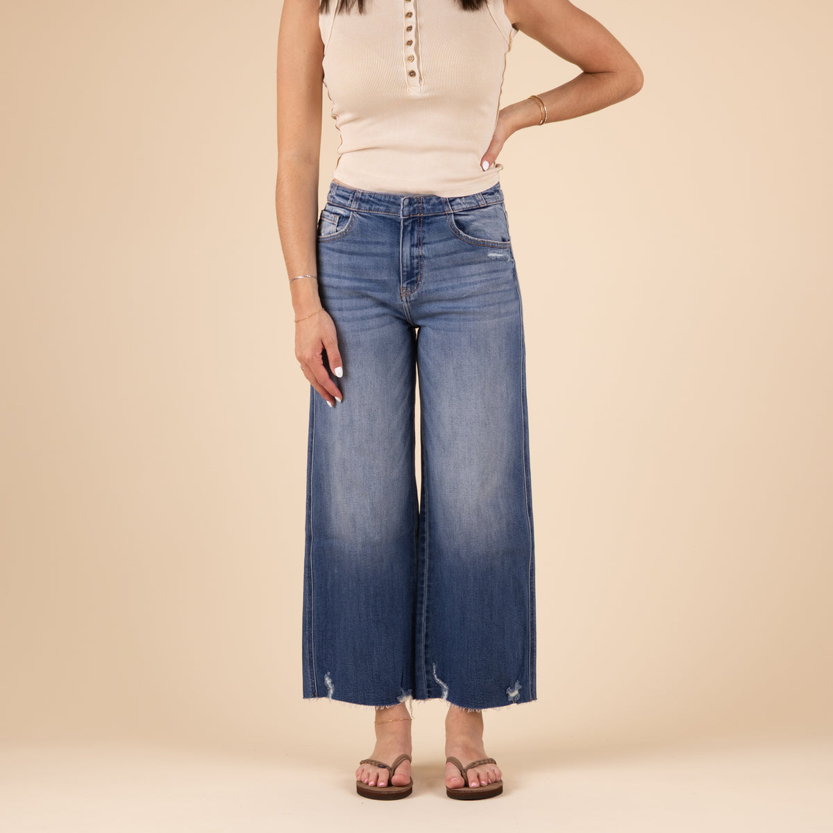 Front view of a woman wearing high-rise, medium-wash wide leg jeans with a faded front, subtle whiskering, light distressing near the hem, and raw edge details. She is also wearing a sleeveless beige top and sandals, standing against a neutral background.
