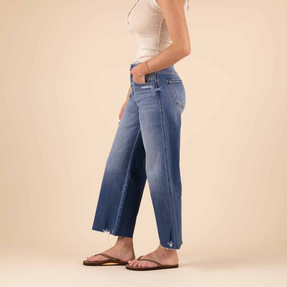 Side view of the wide leg jeans showing the relaxed fit through the hip and thigh, high-rise waist, and slightly flared hem with frayed edges. The model’s beige top and sandals are also visible.