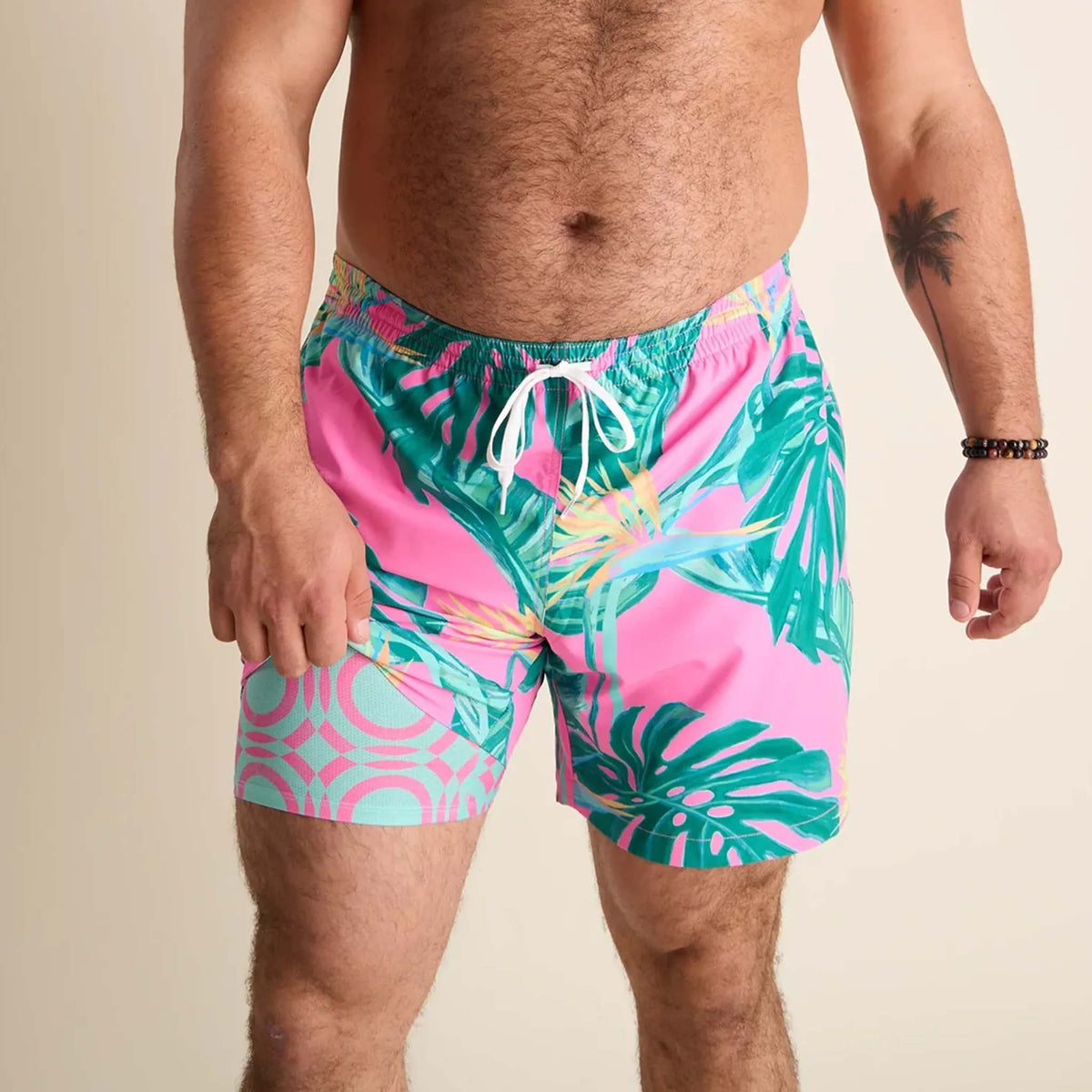 Men’s swim trunks by Chubbies featuring a vibrant tropical design. The shorts have a bright pink background with green palm leaves and tropical foliage printed across the front. They include a white drawstring at the elastic waistband and a built-in liner with a pink and mint green geometric pattern visible near the leg opening.