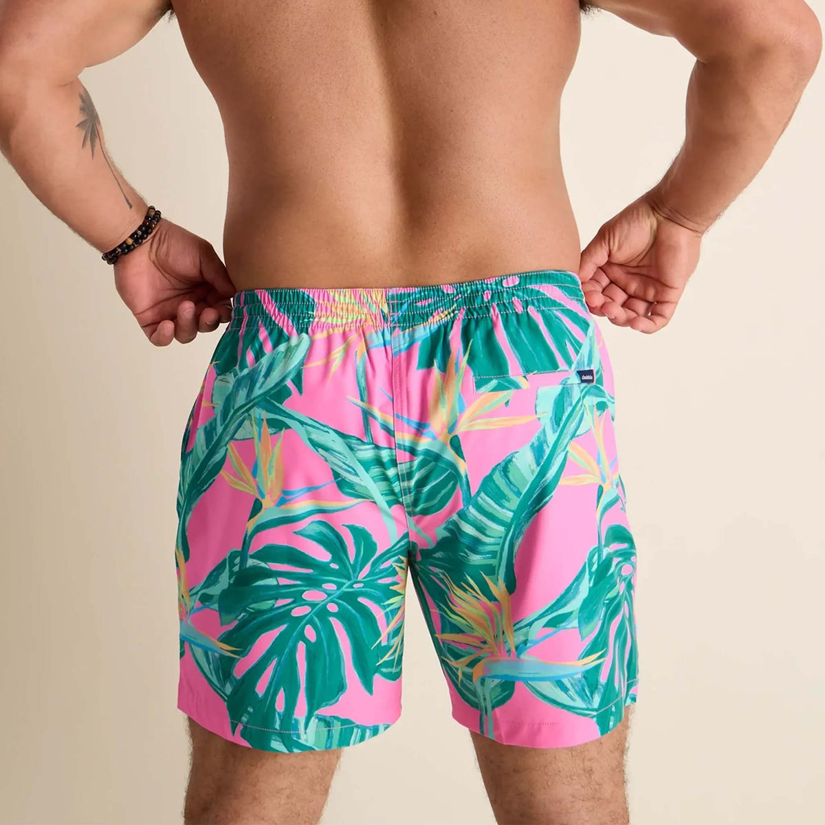 Men’s swim trunks by Chubbies featuring a vibrant tropical design. The shorts have a bright pink background with green palm leaves and tropical foliage printed across the front. They include a white drawstring at the elastic waistband and a built-in liner with a pink and mint green geometric pattern visible near the leg opening.