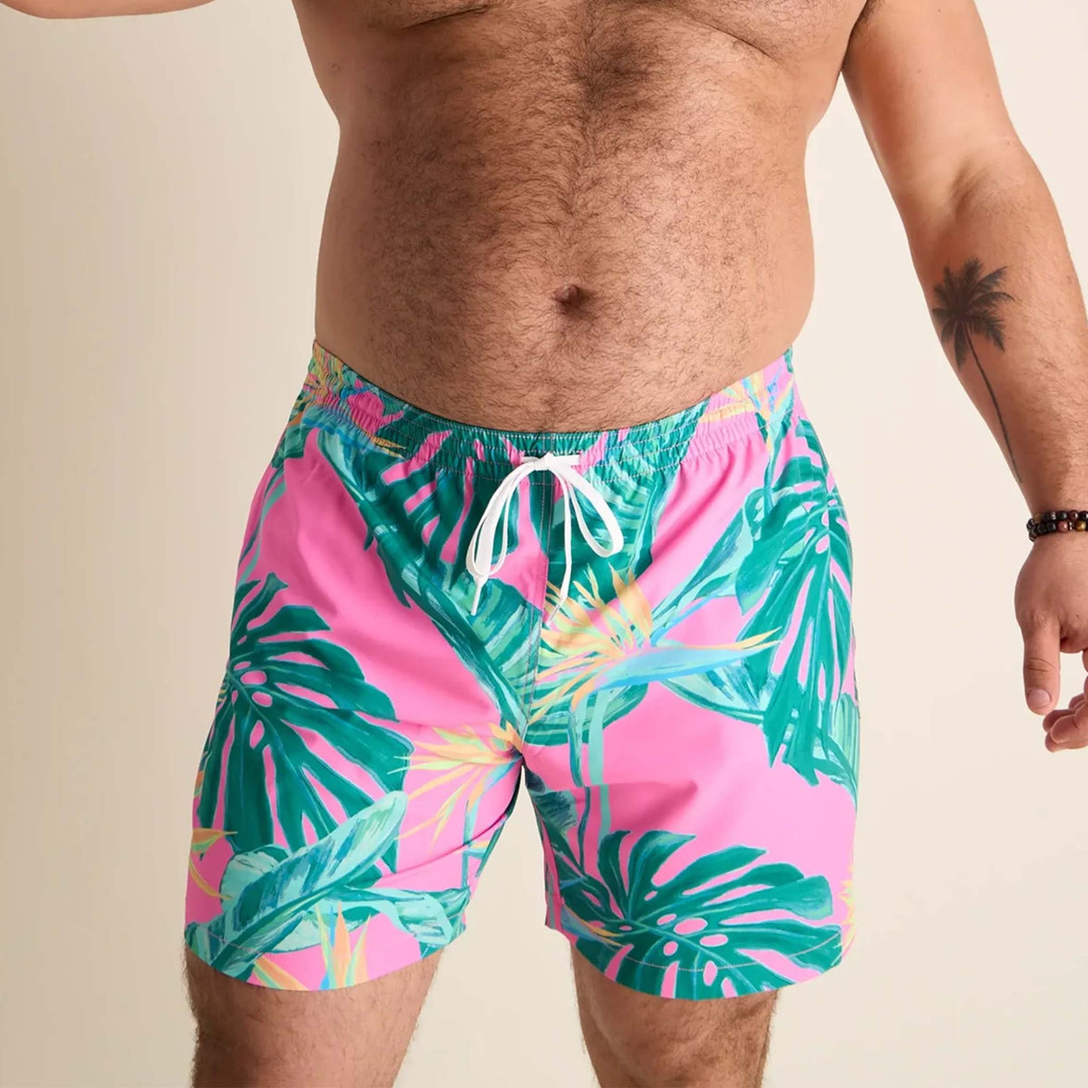 Men’s swim trunks by Chubbies featuring a vibrant tropical design. The shorts have a bright pink background with green palm leaves and tropical foliage printed across the front. They include a white drawstring at the elastic waistband and a built-in liner with a pink and mint green geometric pattern visible near the leg opening.