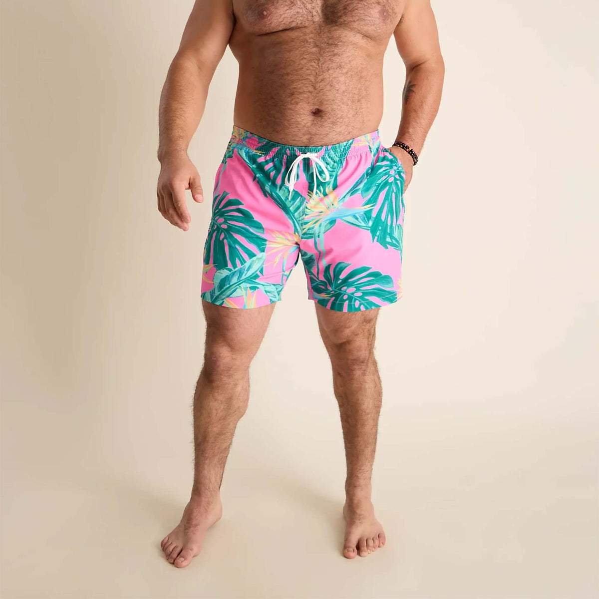 Men’s swim trunks by Chubbies featuring a vibrant tropical design. The shorts have a bright pink background with green palm leaves and tropical foliage printed across the front. They include a white drawstring at the elastic waistband and a built-in liner with a pink and mint green geometric pattern visible near the leg opening.