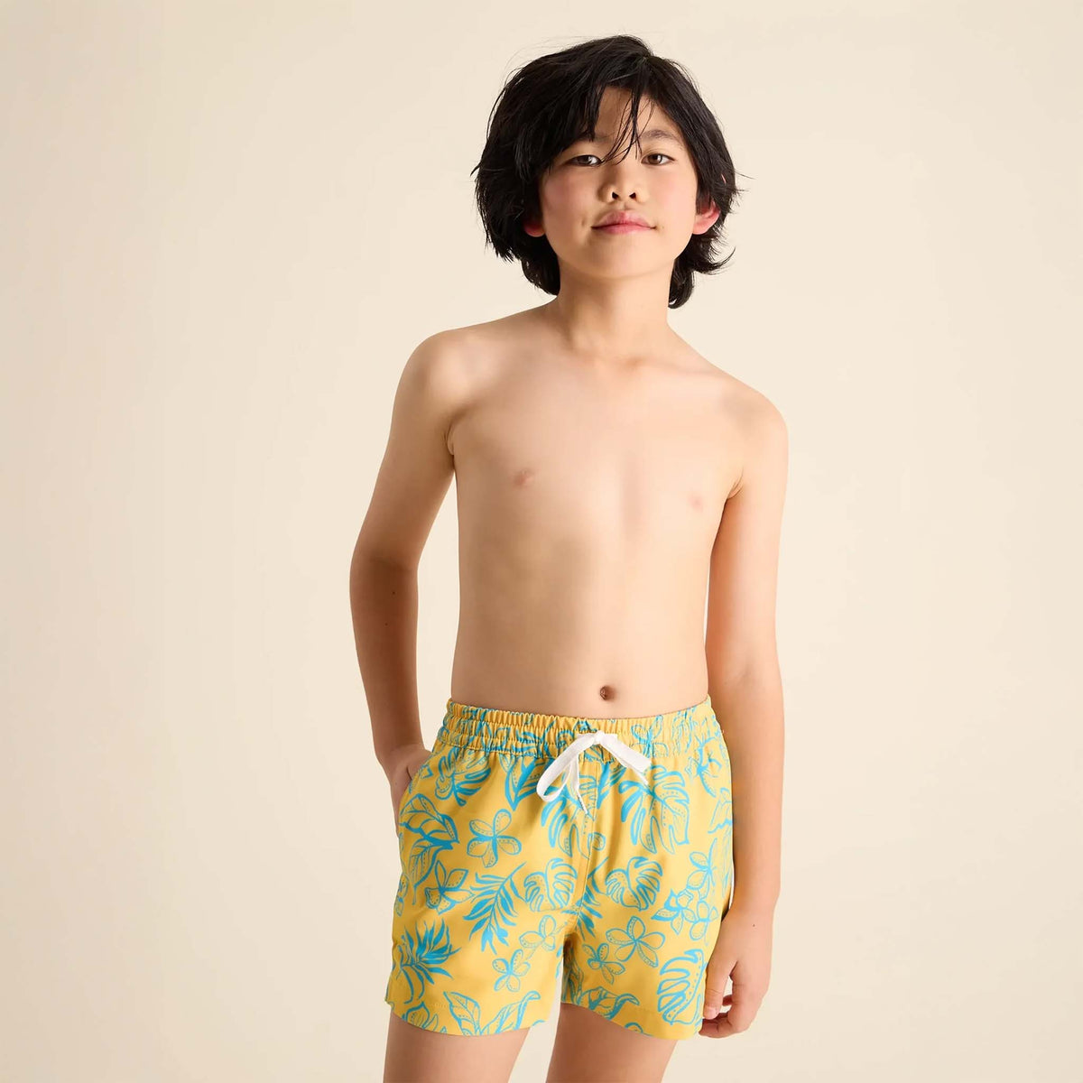 Young boy wearing Youth Lined Swim Shorts in Floral Explorer print by Chubbies, sizes youth S-L.  Print is a warm yellow with sea blue outlines of flowers and monstera leaves throughout.  Liner is sea blue.  Shorts and liner are made with 4-way stretch, polyester/spandex blend.  Drawstring waistband for a comfortable fit, two side pockets, and a zippered back pocket. 