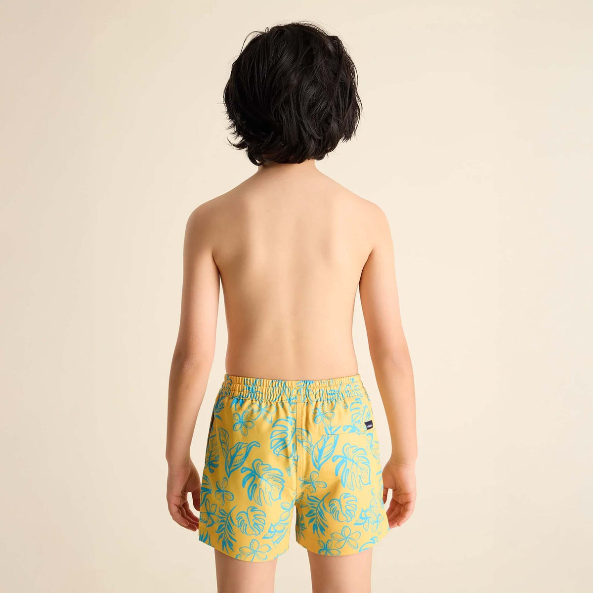 Young boy wearing Youth Lined Swim Shorts in Floral Explorer print by Chubbies, sizes youth S-L.  Print is a warm yellow with sea blue outlines of flowers and monstera leaves throughout.  Liner is sea blue.  Shorts and liner are made with 4-way stretch, polyester/spandex blend.  Drawstring waistband for a comfortable fit, two side pockets, and a zippered back pocket. 
