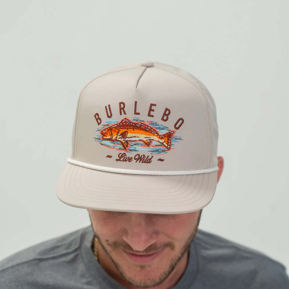 Burlebo rope hat in light khaki with a structured crown and curved bill. The front features an illustration of an orange and red fish over blue water, with the word &#39;BURLEBO&#39; above and the phrase &#39;Live Wild&#39; below. A white rope detail runs across the front panel above the bill. The hat is worn by a man in a gray shirt.