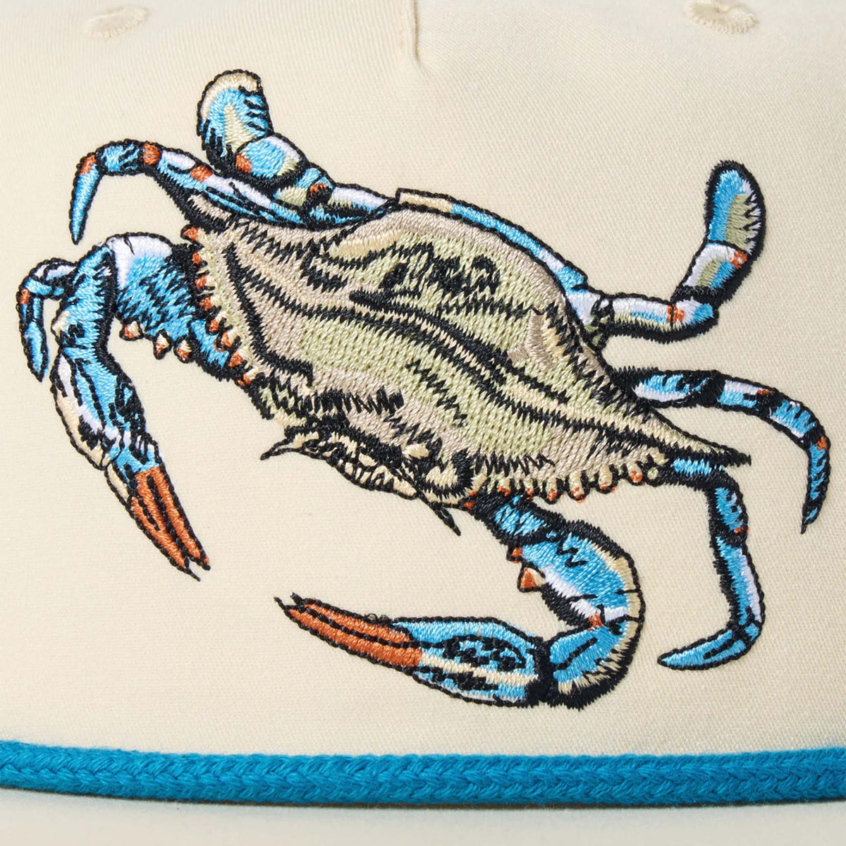 Close-up of the Crab Snapback hat by Duck Camp, color ivory, featuring an embroidered blue crab patch and blue rope cord stretching across the front.  The polyester shell fabric keeps this hat looking good season after season