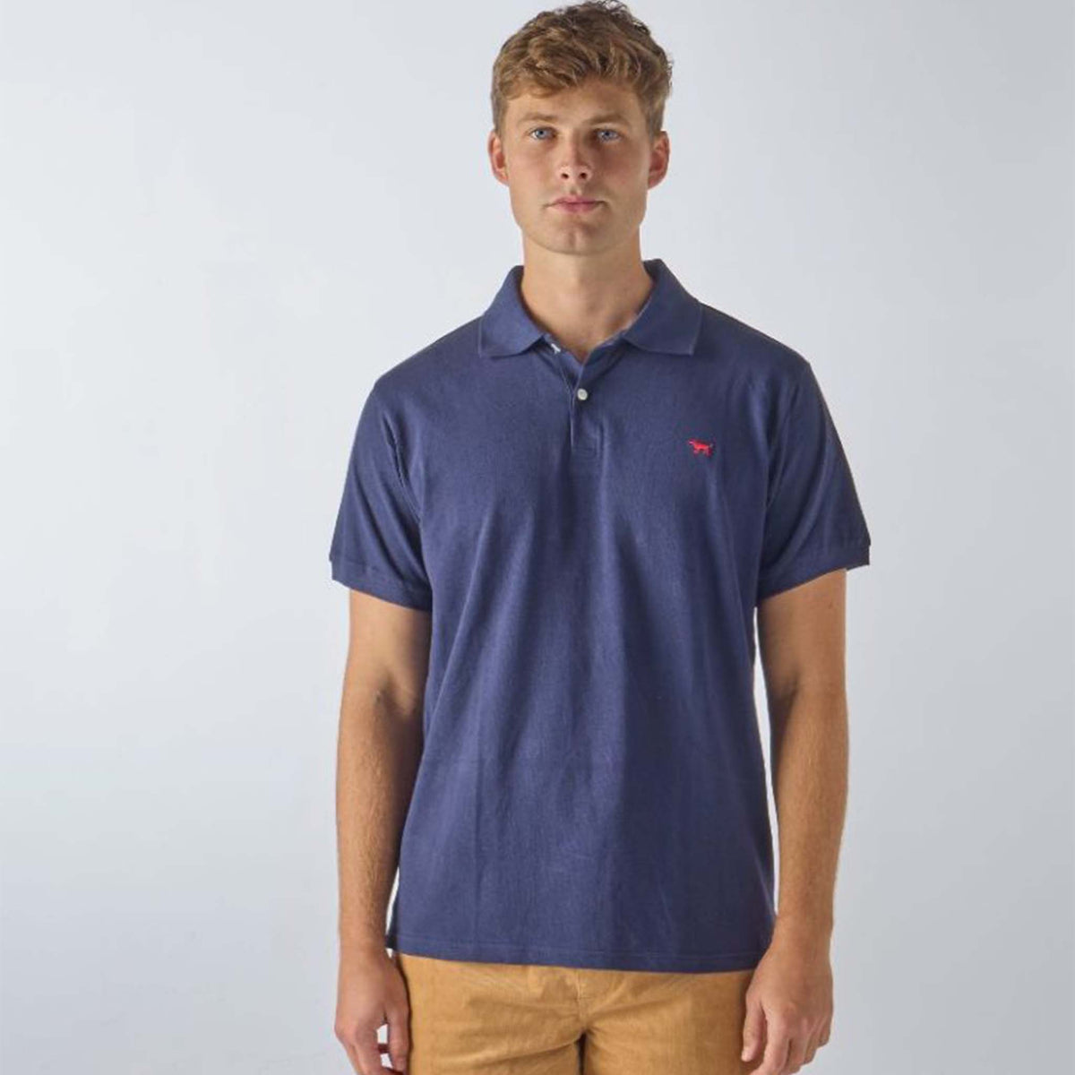 Man wearing a Simply Southern Men&#39;s Solid Polo, color navy, sizes S-XXL, 100% cotton.  There is a 3-button closure at the neckline and a small, embroidered dog embroidered on the left chest. Its classic design features a clean, simple look that&#39;s perfect for both casual and semi-formal occasions. With its versatile style and durable fabric, this polo offers a timeless wardrobe staple for effortless, all-day wear.