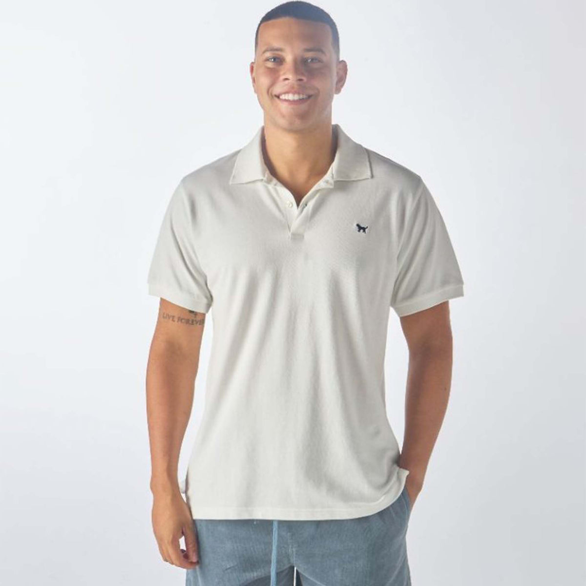 Man wearing a Simply Southern Men&#39;s Solid Polo, color white, sizes S-XXL, 100% cotton.  There is a 3-button closure at the neckline and a small, embroidered dog embroidered on the left chest. Its classic design features a clean, simple look that&#39;s perfect for both casual and semi-formal occasions. With its versatile style and durable fabric, this polo offers a timeless wardrobe staple for effortless, all-day wear.